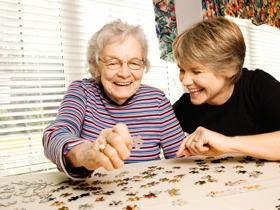 Eine jüngere Frau unterstützt eine alte Dame beim Puzzeln. Beide lachen.